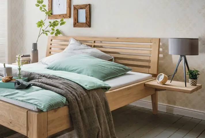 Bett aus hellem Holz mit Lamellenkopfteil, grüner Bettwäsche und grauer Decke, rechts schwebende Ablage mit Tischlampe und kleinem Pflanztopf.